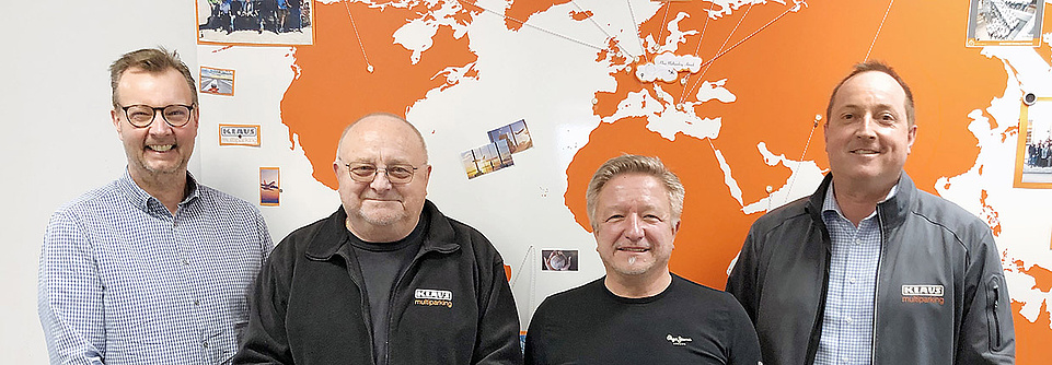 Vier Männer schauen nach vorne, im Hintergrund eine Weltkarte in orange