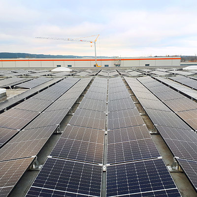 Reihen von Photovoltaik-Anlagen auf einem Dach, im Hintergrund Firmengebäude und Himmel