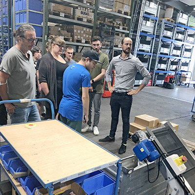 There are several people in a storage area, with a high wall of shelves in the background