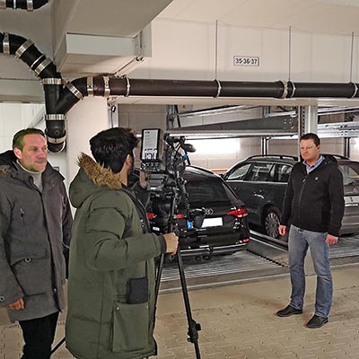 Two men are conducting an interview in front of parked cars, and a cameraman is filming