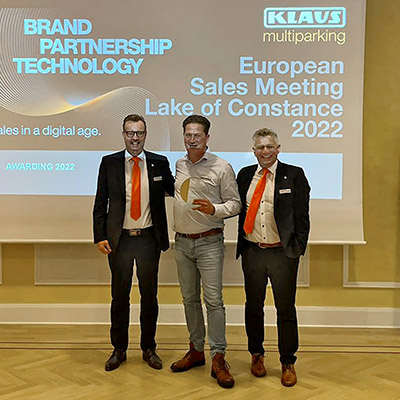 Three men in front of a projection screen, the man in the middle is holding a trophy