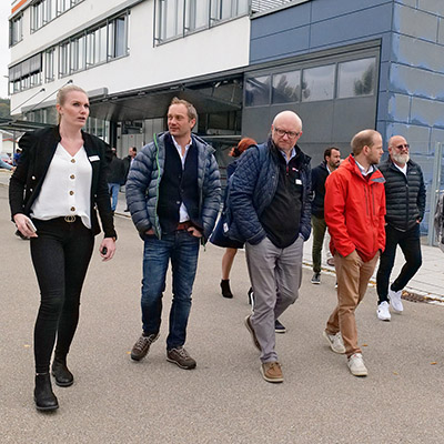 Mehrere Personen laufen vor einem Firmengebäude, besichtigen und reden miteinander